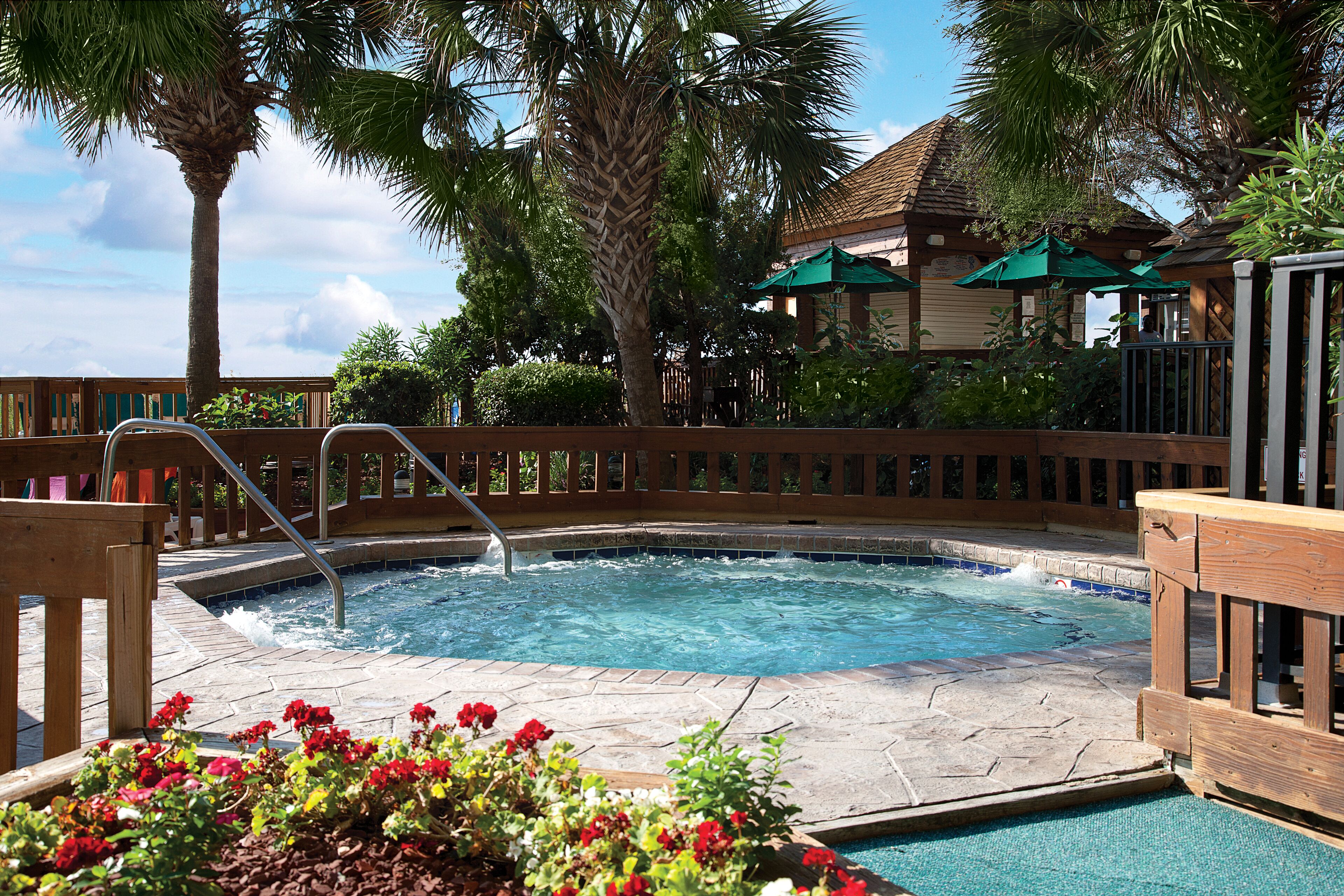 A hot tub area with a wooden deck, a pool, and a building with green umbrellas in the background.