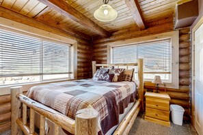 A log cabin bedroom with a wooden bed, a nightstand, and a window with blinds. - Beautiful log cabin with lake views, wraparound deck, & foosball (Garden City)