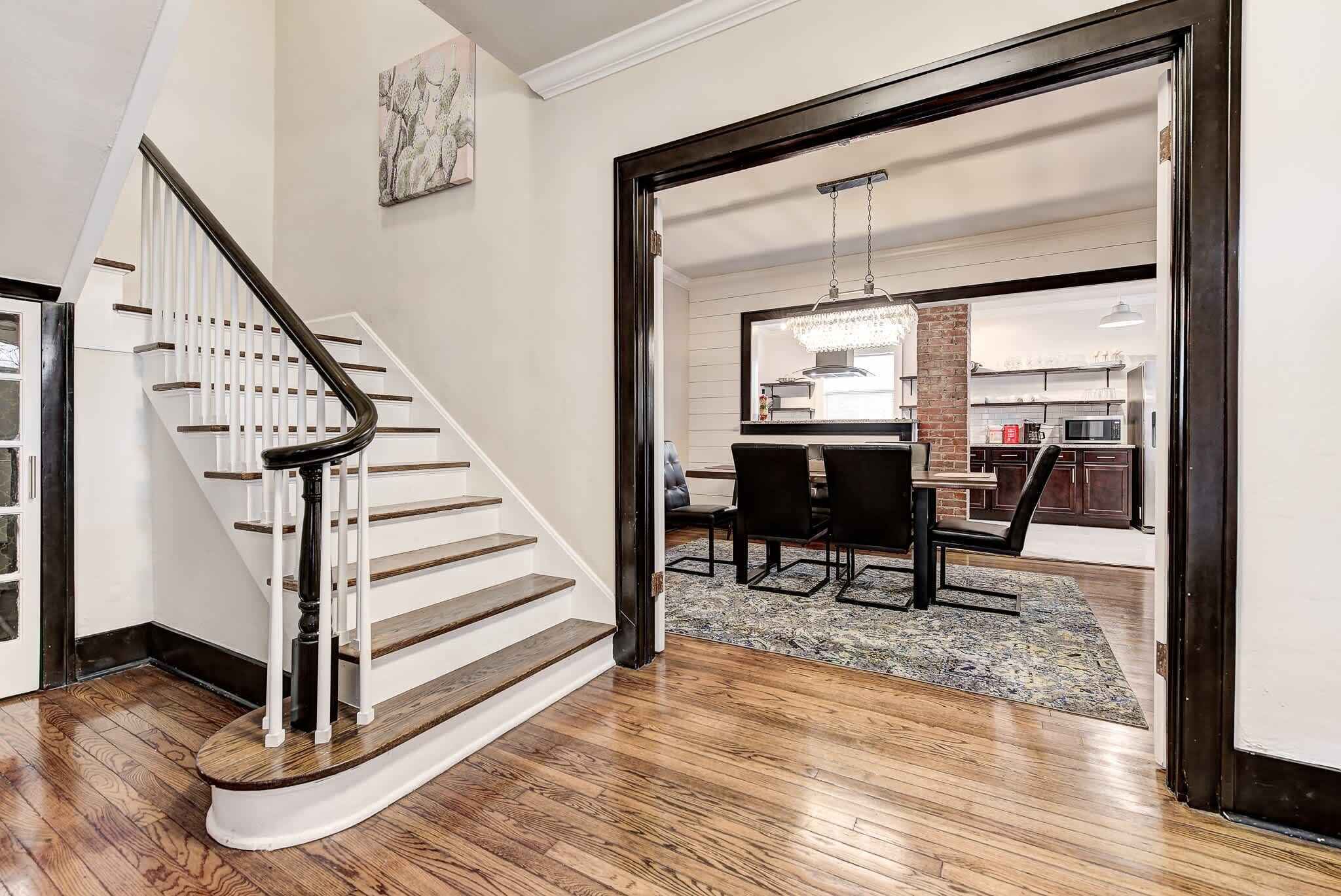 A staircase leading to a dining area with a chandelier, a kitchen in the background, and a rug on the floor.