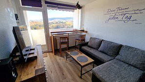 A cozy living room with a sofa, a coffee table, and a dining table with chairs. A large window offers a view of a forested landscape.