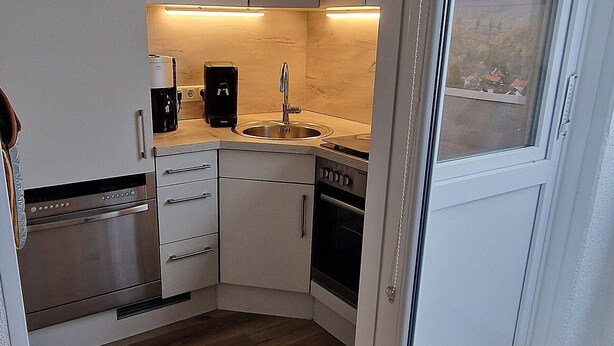 A compact kitchen with white cabinets, a built-in oven, a coffee maker, and a window with a view of a hilly landscape.