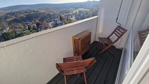 A balcony with a wooden bench and a small wooden chest, overlooking a village and hills.