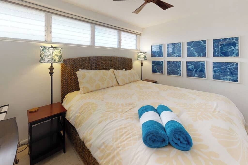 A hotel room with a bed, two bedside lamps, a ceiling fan, a window with blinds, and six framed pictures on the wall.