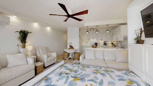 A modern, well-lit room with a ceiling fan, a sofa, a small table with a lamp, and a kitchen area visible through an open door. - Perfect location and Unbelievable Views at Lahaina Roads (Lahaina)