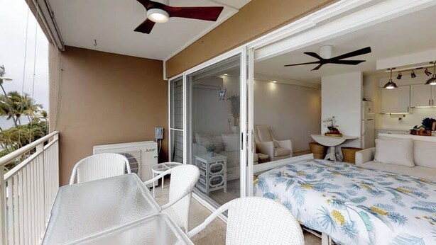 A balcony with a table and chairs, a sliding glass door leading to a bedroom with a bed and a small table, and a ceiling fan. - Perfect location and Unbelievable Views at Lahaina Roads (Lahaina)