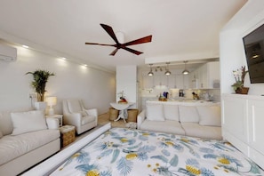 A modern, well-lit room with a ceiling fan, a sofa, a small table with a lamp, and a kitchen area visible through an open door.