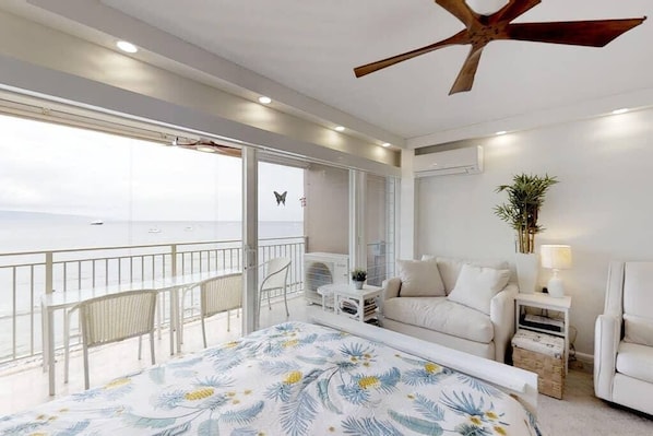 A balcony with a view of the sea, a bed with floral bedding, a white sofa, a small table, and a ceiling fan.