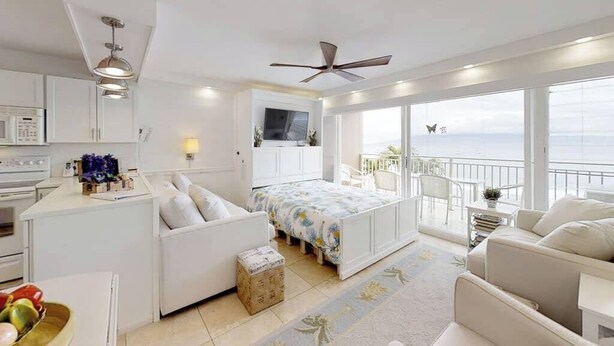 A modern bedroom with a balcony, a bed with floral bedding, a white sofa, and a kitchen area with a microwave. - Perfect location and Unbelievable Views at Lahaina Roads (Lahaina)