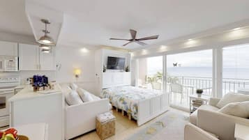 A modern bedroom with a balcony, a bed with floral bedding, a white sofa, and a kitchen area with a microwave.