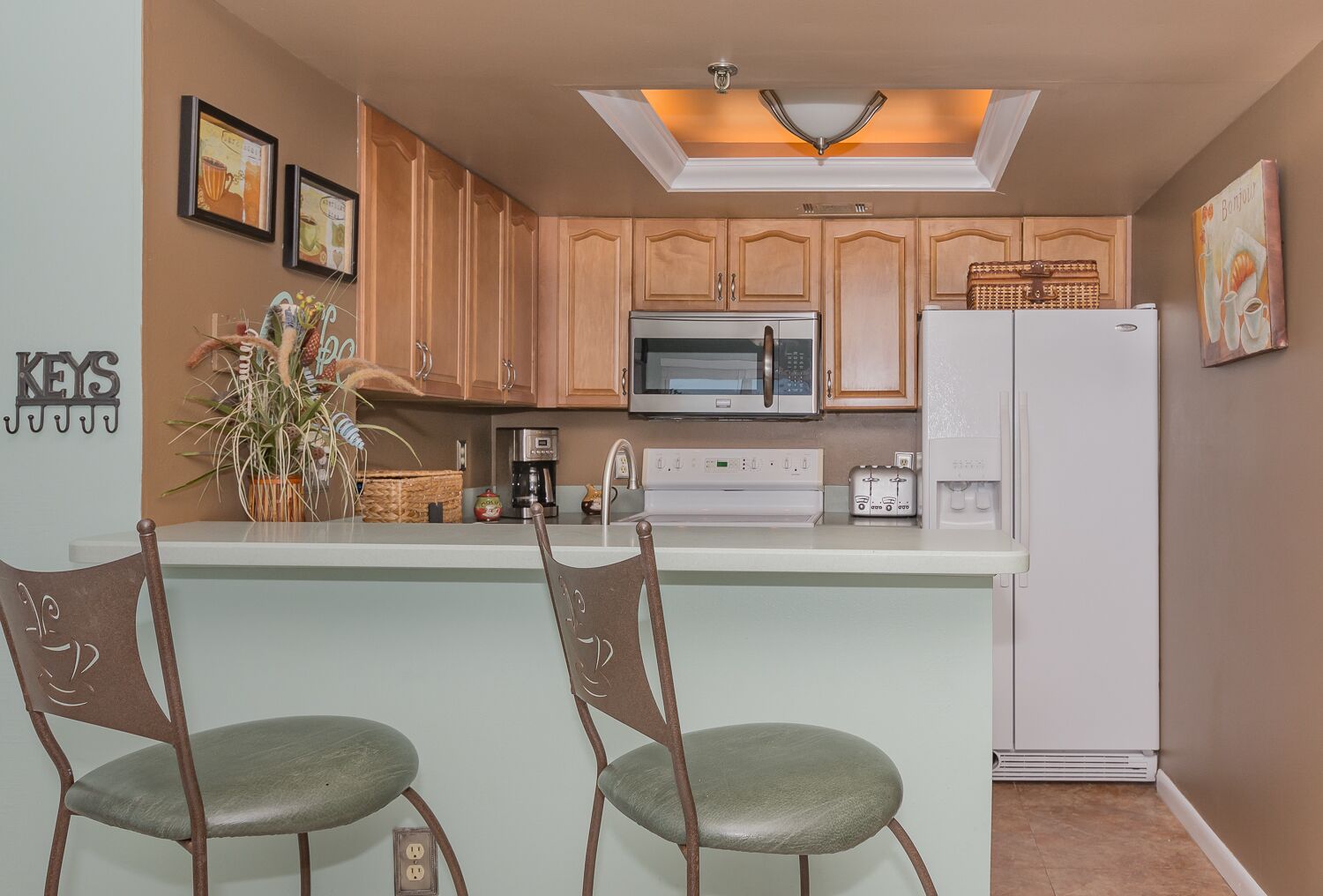 A kitchen with wooden cabinets, a microwave, a refrigerator, and a bar area with two chairs.