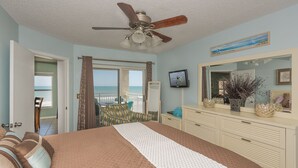 A bedroom with a bed, a ceiling fan, a television, a dresser, and a view of the ocean through a sliding door.