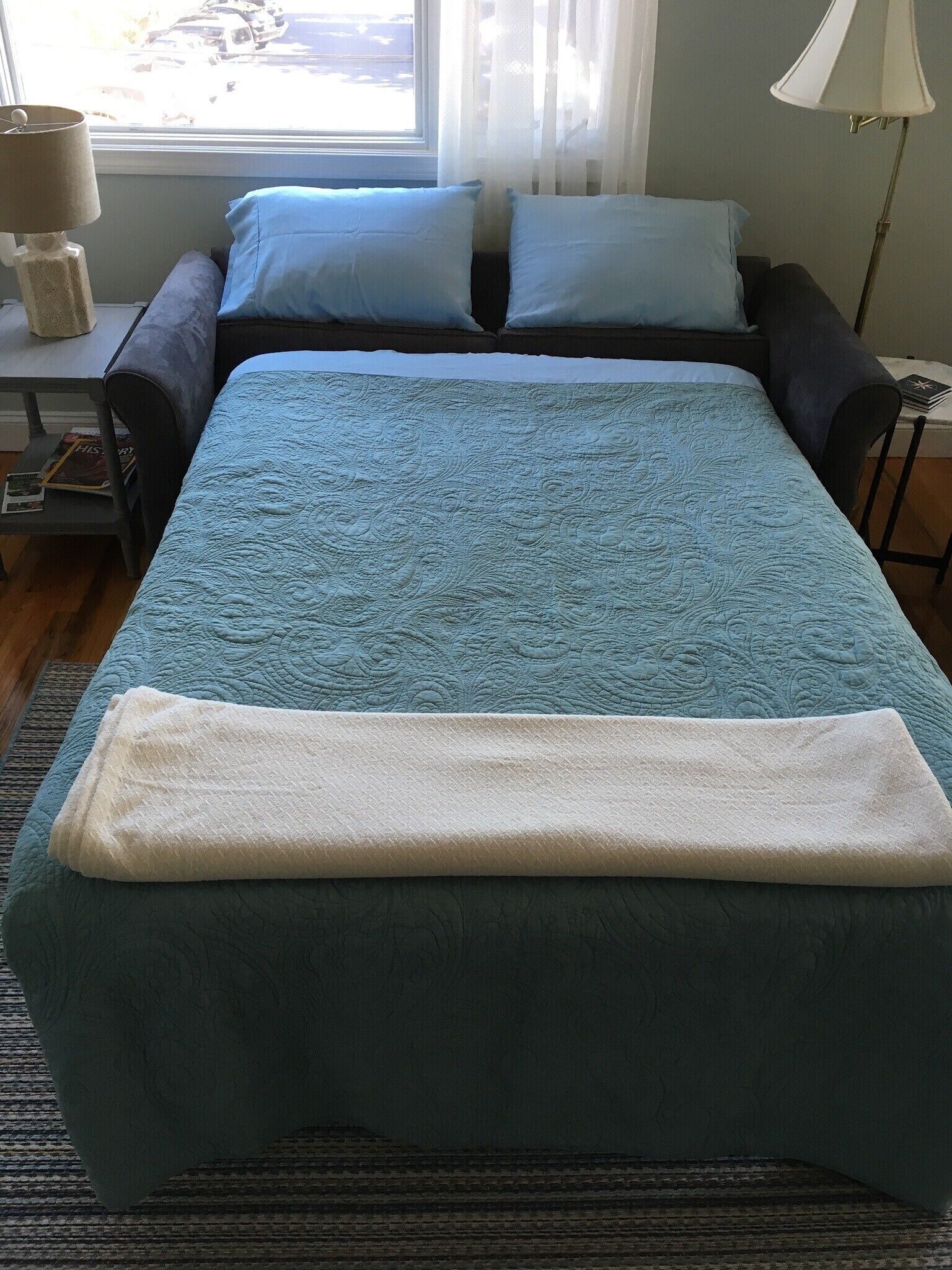 A neatly made bed with a blue quilt and a white blanket, a small table with a lamp, and a window with sheer curtains.