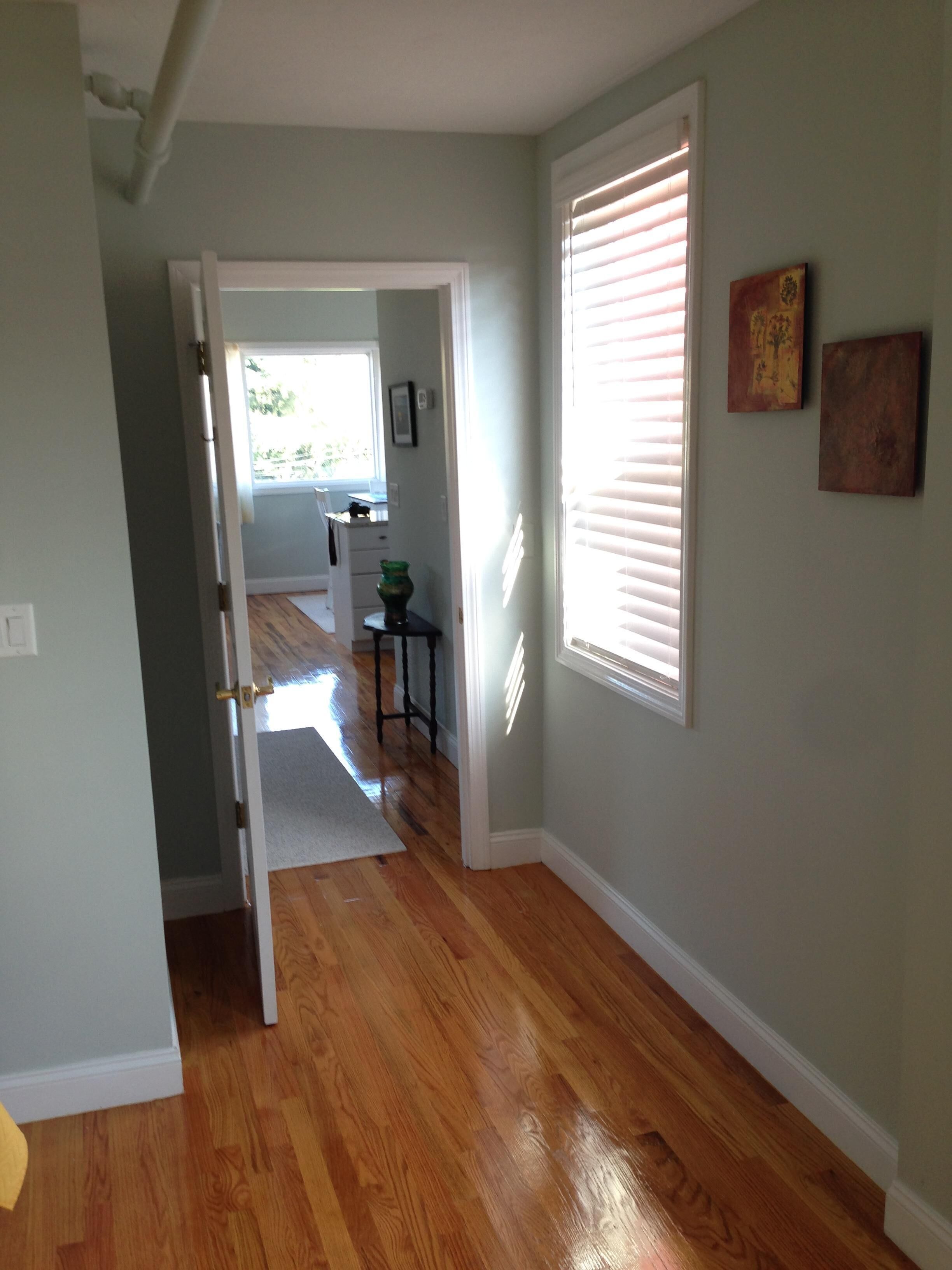 A hallway with wooden flooring, a door leading to another room, and a window with blinds.
