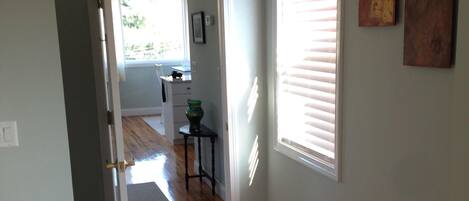 A hallway with wooden flooring, a door leading to another room, and a window with blinds.