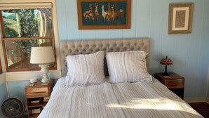 A bedroom with a tufted headboard, striped bedding, a window with a view of trees, a bedside table with a lamp, and a framed picture on the wall. - Family and pet friendly close to beaches. SC Rental Permit 241396 (Santa Cruz)
