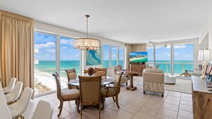 A spacious room with a large window overlooking the beach, a dining area with chairs and a table, and a view of the ocean. - Beautiful Condo with Amazing Views, Beach Service (Navarre)