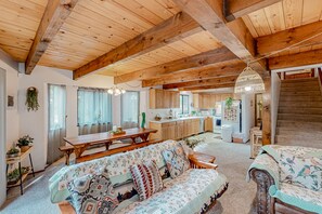 A spacious living area with a wooden ceiling, a sofa with patterned cushions, a dining table, and a kitchen in the background.