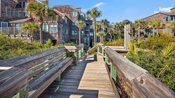 A wooden boardwalk leading to a building with brick walls and multiple windows, surrounded by palm trees and greenery. - Windswept 4463! Oceanfront 3 BR Condo, Great Location! (Kiawah Island)