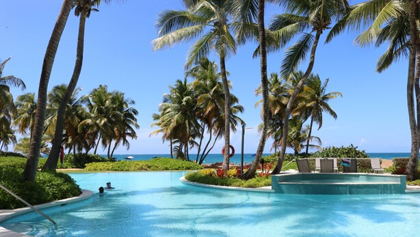 A pool area with palm trees, a clear sky, and a view of the ocean. -  Beautiful beachfront villa at the Wyndham Grand R Mar Rainforest Beach Resort  (Rio Grande)