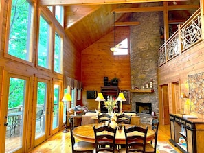 Interior acogedor de una cabaña con chimenea de piedra, una mesa de comedor de madera y ventanas amplias.