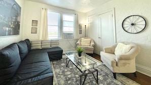 A living room with a sectional sofa, a glass coffee table, a white armchair, and a dartboard on the wall. - 6653 2S | Jewel of Central St. Louis (St. Louis)