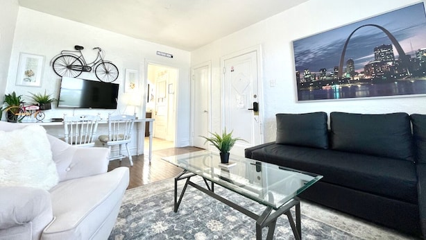 A living room with a white sofa, a black sofa, a glass coffee table, and a cityscape painting. - 6653 2S | Jewel of Central St. Louis (St. Louis)