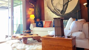 A cozy living room with a wooden ceiling, a large abstract painting, a white sofa with colorful cushions, a wooden coffee table, and a lamp.