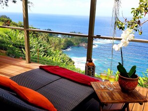 Un balcon doté d’un plancher en bois, un canapé avec des coussins orange, une petite table sur laquelle est posée une plante en pot et deux verres remplis de boisson, et une lanterne suspendue.