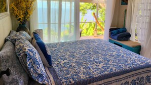 A bedroom with a bed, pillows, a vase of yellow flowers, and a view of greenery outside.