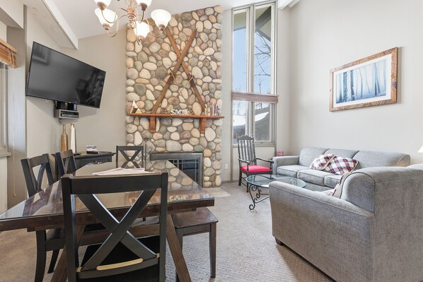 A living room with a stone wall, a fireplace, a flat-screen TV, a dining table with chairs, and a grey sofa.