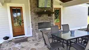 Une terrasse extérieure avec un sol en pierre, une cheminée en pierre, un téléviseur fixé au mur et une table avec des chaises.