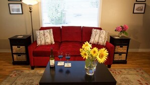 A red sofa with floral cushions, a coffee table with a vase of sunflowers, a bottle of wine, and two glasses. - Syvila’s Cottage | Private Garden Near Downtown (Walla Walla)