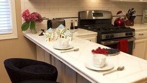 A kitchen with a white countertop, a stove, a toaster, and a red kettle. There are two chairs and a vase with flowers on the counter. - Syvila’s Cottage | Private Garden Near Downtown (Walla Walla)