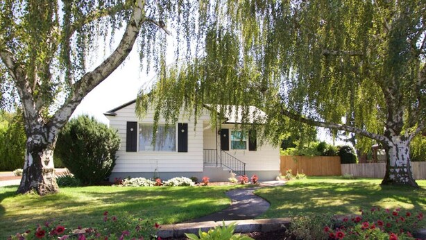 A single-story house with a front porch, surrounded by a well-maintained lawn and flowering plants. - Syvila’s Cottage | Private Garden Near Downtown (Walla Walla)