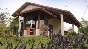 Ein Haus mit Terrasse, einem Kamin aus Ziegeln und einem Garten mit violetten Blumen.