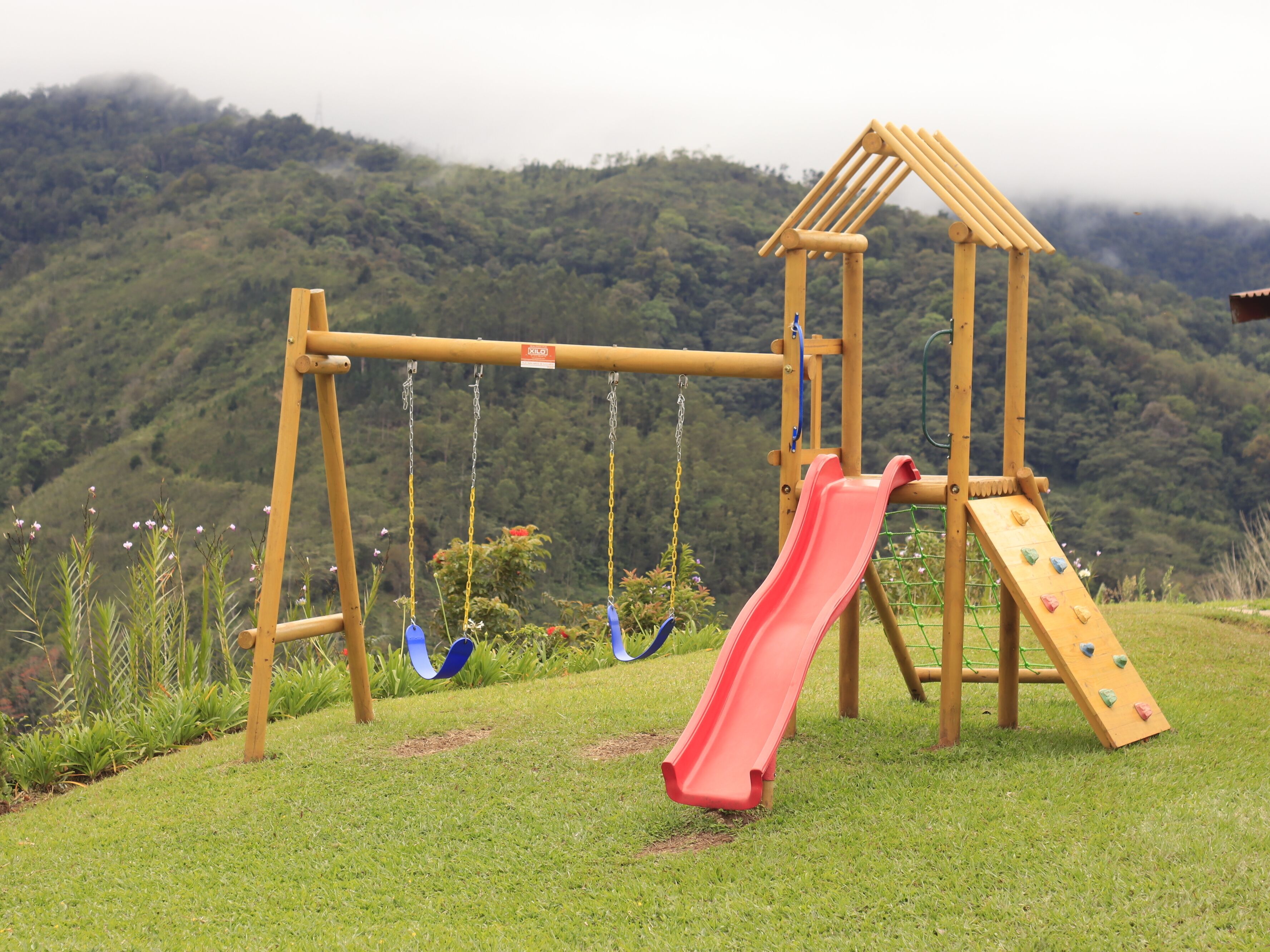 A wooden playground with a slide and swings, set against a backdrop of a forested hill.
