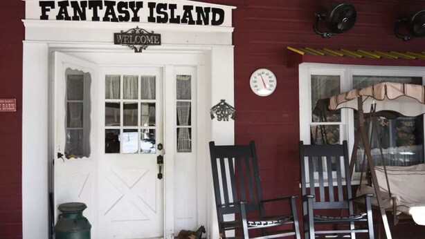 Una puerta con la leyenda 'Fantasy Island', un cartel de 'Bienvenido' arriba, dos mecedoras adelante y un reloj en la pared.