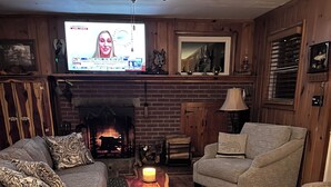 Un salon chaleureux avec une cheminée, des fauteuils confortables et un téléviseur diffusant un programme d’information.
