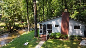 Une petite maison de plain-pied, avec une cheminée en briques, entourée d’une végétation luxuriante et bordée d’un ruisseau.