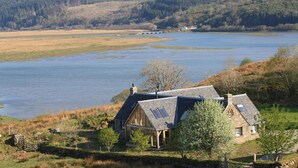 Una casa con techo inclinado y ventanas amplias, ubicada cerca de un lago con una colina boscosa al fondo.
