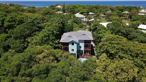 Uma casa de dois andares rodeada por vegetação tropical exuberante, com vista para o oceano ao fundo.