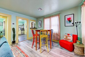 A room with a dining table, chairs, a sofa, a small side table, and a kitchen in the background.