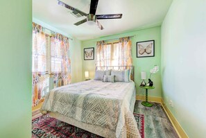 Un dormitorio con cama, cortinas, ventilador de techo, mesita de noche, lámpara y una pequeña mesa con una orquídea.