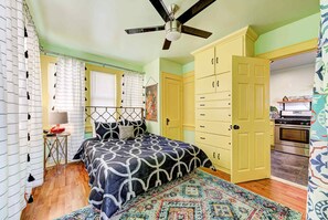 Un dormitorio con cama, ventilador de techo, armario, ventana con persianas y una zona de cocina.