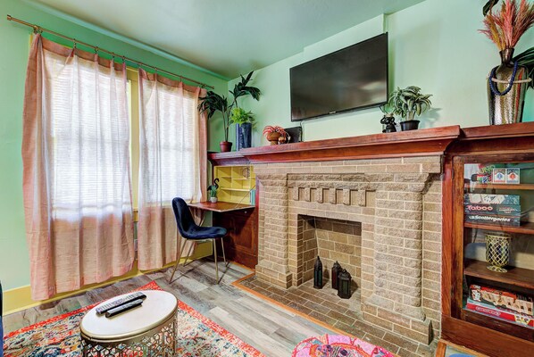 Una sala de estar con chimenea de ladrillo, un televisor montado encima, un sillón azul y una estantería con juegos de mesa.