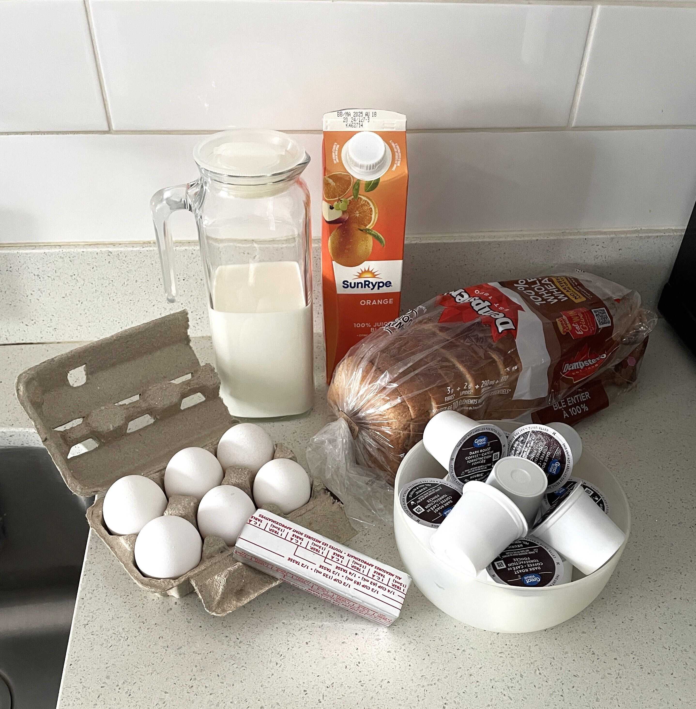 A kitchen counter with a carton of orange juice, a glass pitcher of milk, a loaf of bread, a carton of eggs, and several coffee pods.