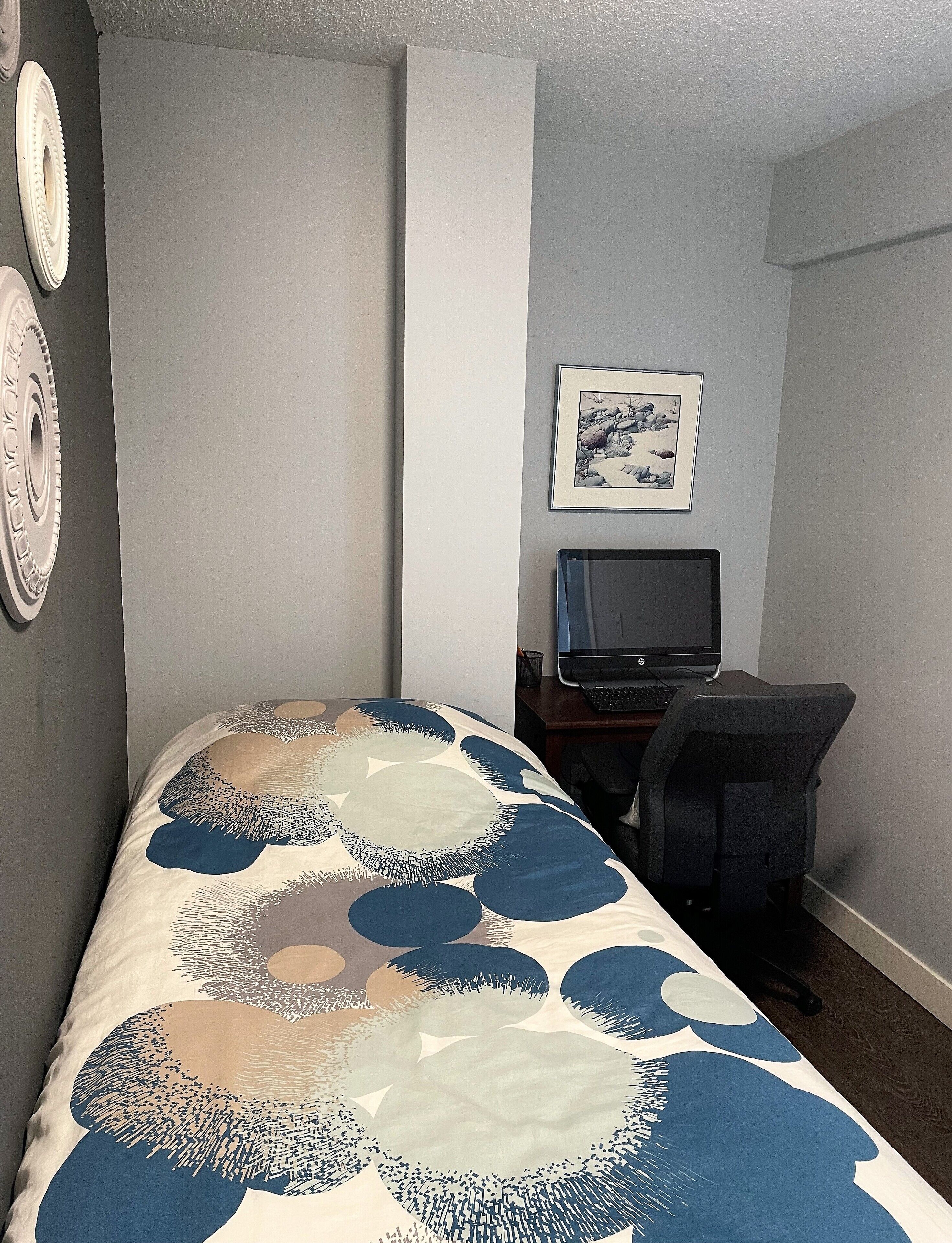 A bedroom with a bed, a desk, a chair, a computer, and a framed picture on the wall.