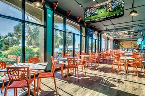 Un restaurante espacioso con amplias ventanas, sillas anaranjadas y una pantalla de televisión mostrando un partido deportivo.