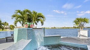 A pool area with a waterfall feature, surrounded by palm trees and a view of the water body.