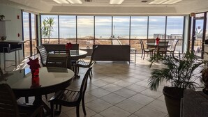 A spacious indoor area with a large window overlooking a beach, furnished with tables and chairs, and a potted plant. - PHOENIX V..GULF FRONT..10' CEILINGS..WKLY DISCOUNTS..RESERVED COVERED PARKING. (Orange Beach)